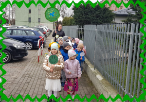 Na zdjęciu grupa dzieci przed przedszkolem. . Każde dziecko trzyma plakietkę na patyczku.